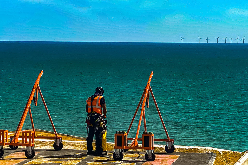 Construction worker on seaside platform with orange equipment, gazing at distant wind turbines on ocean horizon.