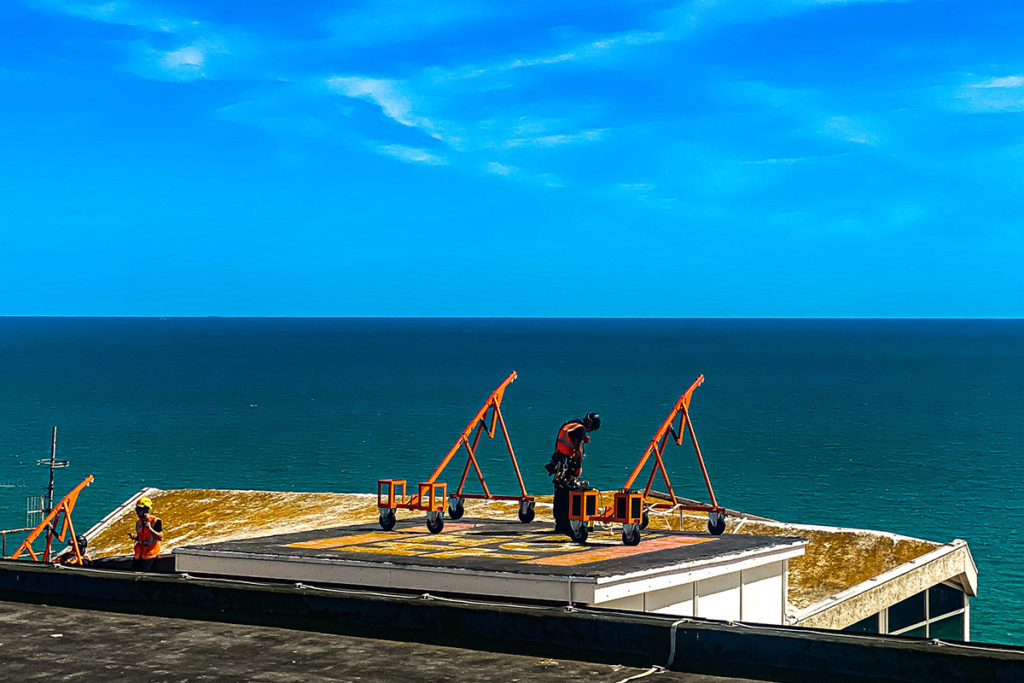 Workers in orange vests operating equipment on a rooftop, overlooking the deep blue sea under a clear sky.