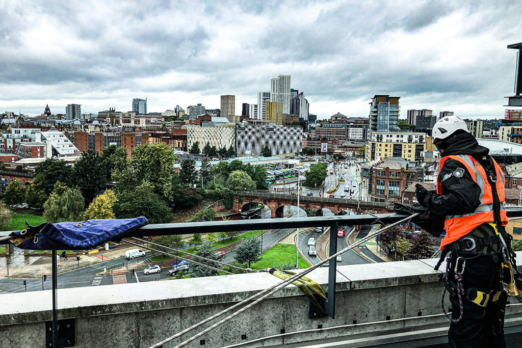 Worker in safety gear on rooftop overlooking Leeds cityscape with cloudy sky. Urban landscape, high angle.