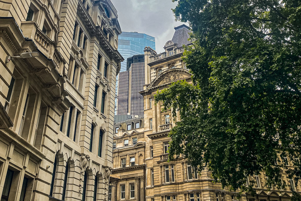 Historic stone buildings with ornate facades beside modern skyscrapers under a cloudy sky, framed by trees.