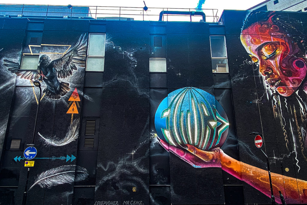 Colourful street art mural on dark wall, featuring a bird, abstract face, and hand holding a geometric orb.