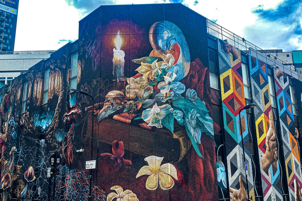Colourful street mural featuring candles, flowers, and geometric patterns on building façade, vibrant skies above.