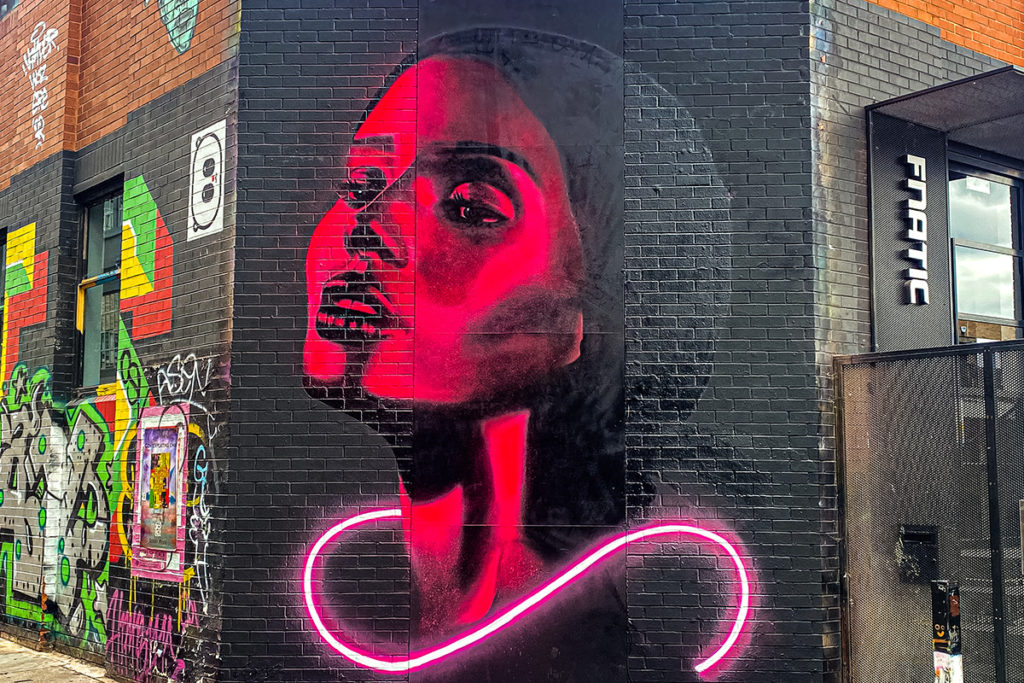 Vivid pink and black mural of a woman's face on a brick wall beside FNATIC store, with neon accents.