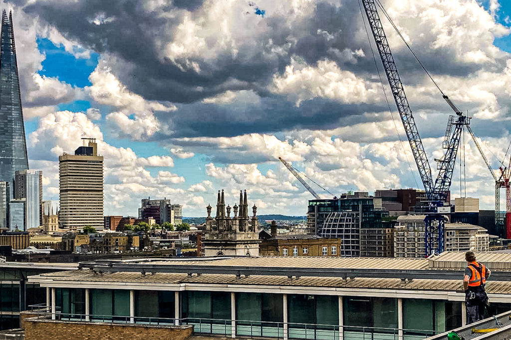 London skyline with the Shard, cranes, and dramatic clouds; worker in orange vest on rooftop in foreground.