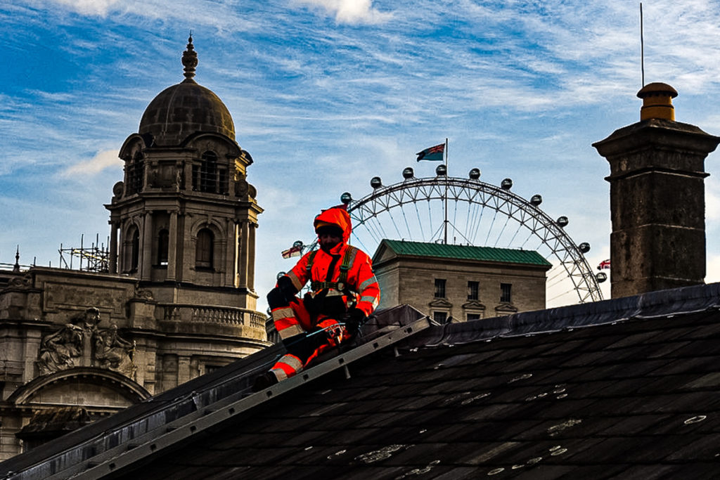 Worker in high-visibility gear on a rooftop in London, with the London Eye and historic buildings visible.