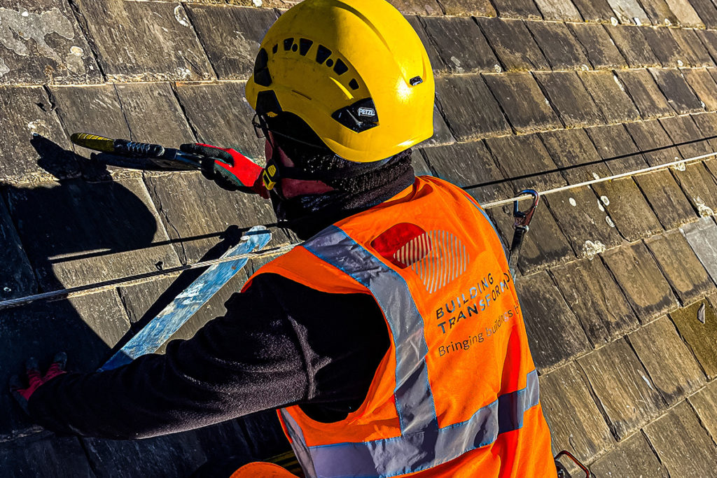 Roofer in yellow helmet and orange vest works on slate roof with safety gear; Building Transformation.