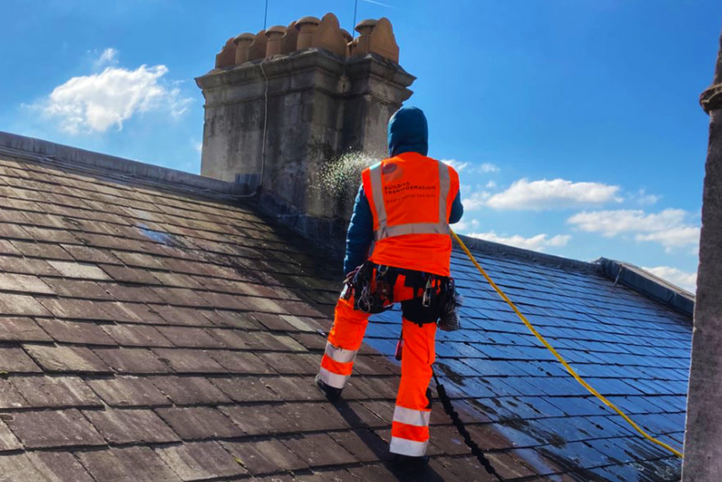 Worker in orange safety gear power-washing a slate roof on a sunny day, blue sky in background.
