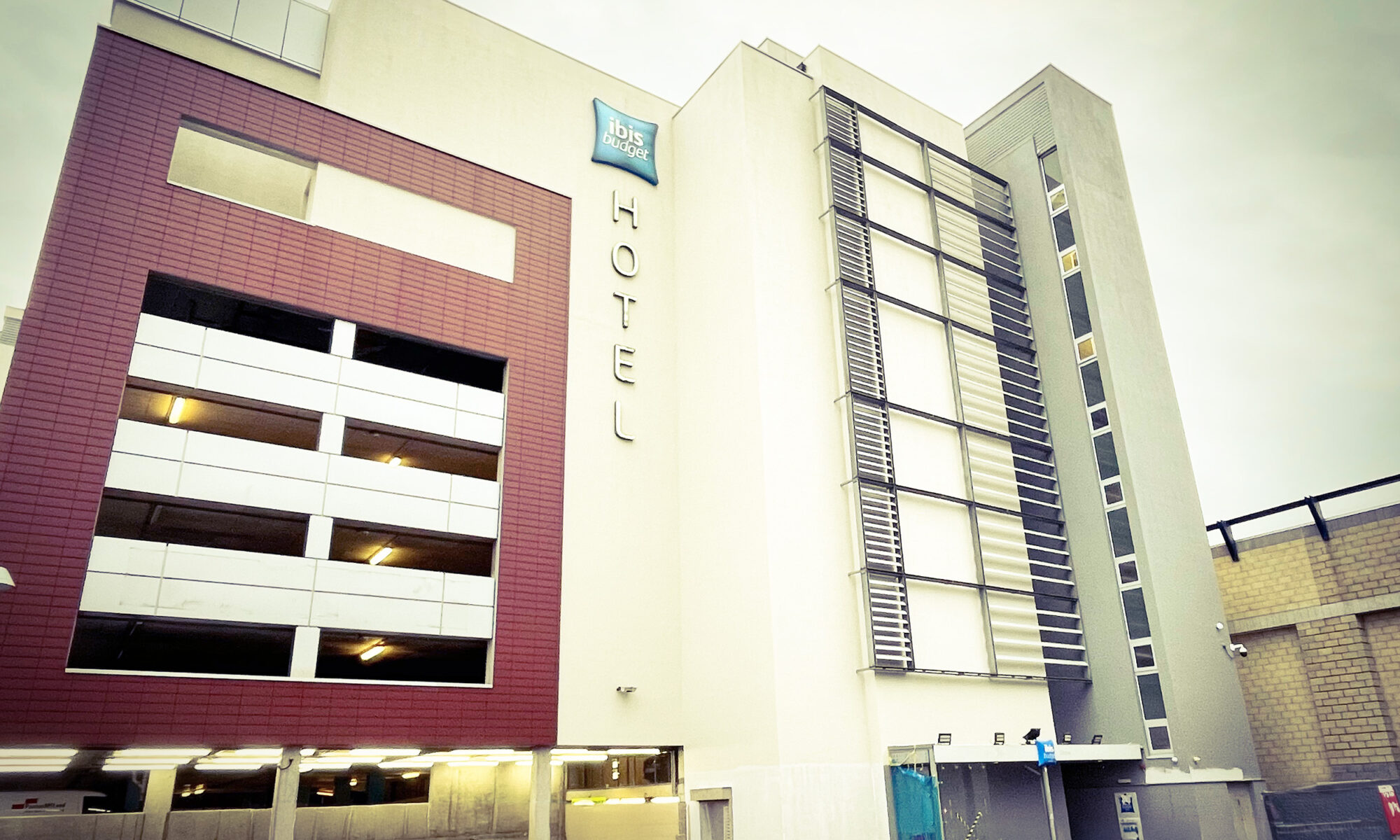 Modern Ibis Budget Hotel exterior with vertical HOTEL sign and garage entrance on cloudy day.
