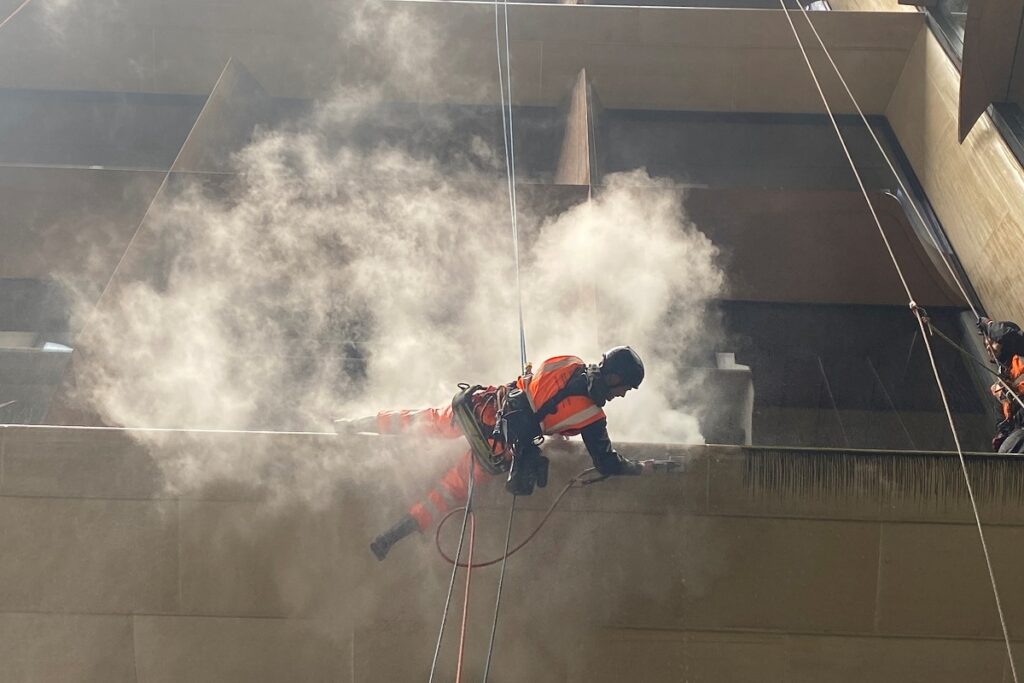 Worker in bright orange safety gear abseiling down a tall building, surrounded by concrete dust.