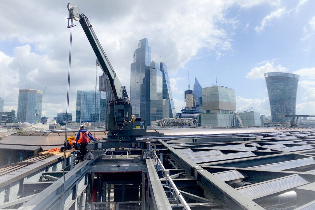Construction workers on a steel roof with crane, London skyscrapers in background under a partly cloudy sky.