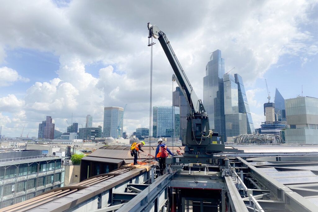 Industrial workers in safety gear operate a crane on a rooftop, with London's skyline in the background.