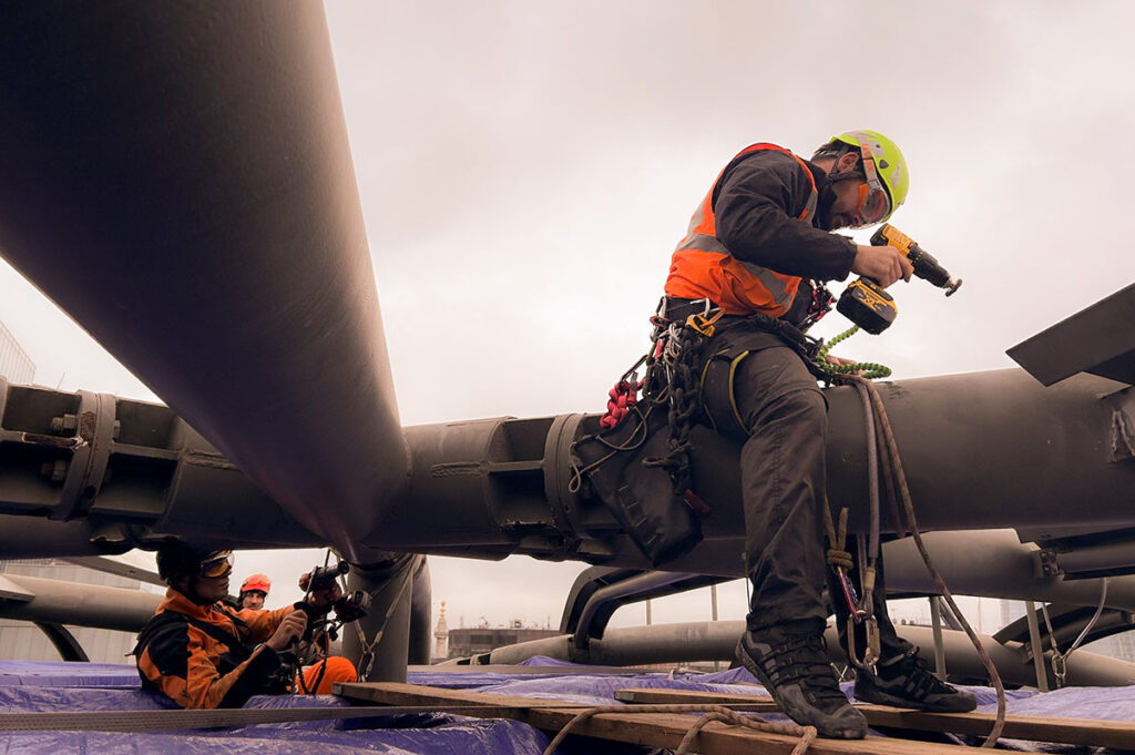 Construction workers in safety gear, using power tools on large grey pipes under a cloudy sky.
