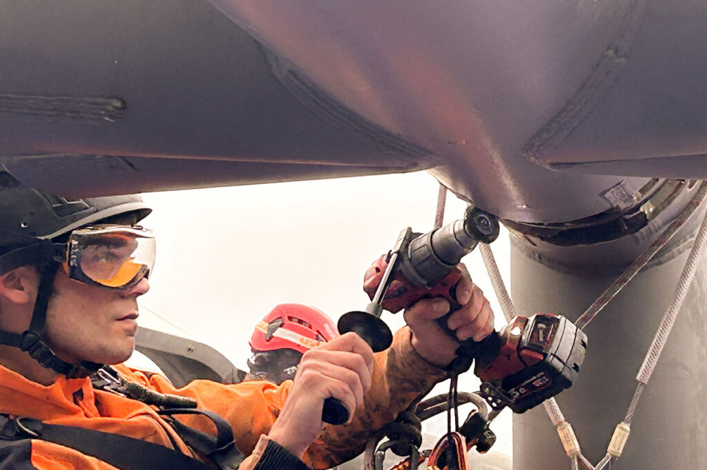 Worker in orange gear and safety helmet uses a power drill on a large metal structure, focused and alert.