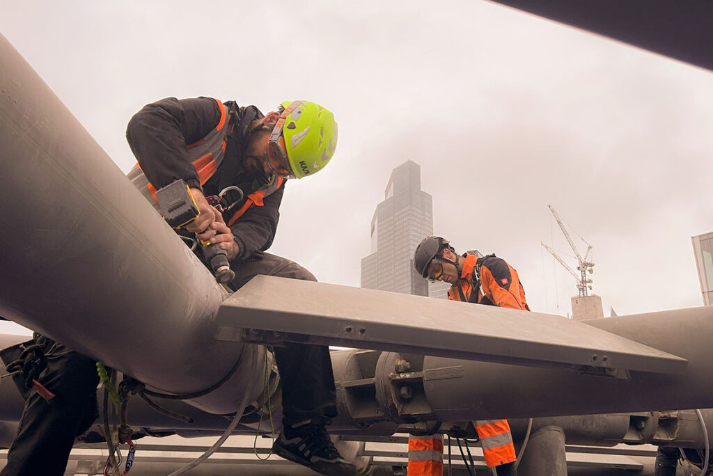 Industrial workers in safety gear using power tools on a rooftop, urban skyline and cranes in background.