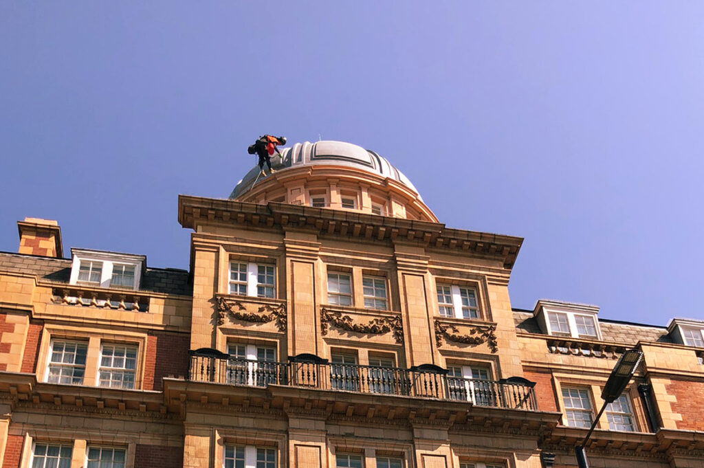 Worker repairing rooftop dome on a historic building under clear blue sky, showcasing architectural details.