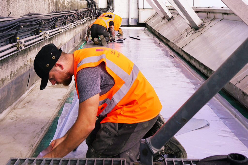 Construction workers in orange vests kneeling on a rooftop, sealing a surface with protective material.