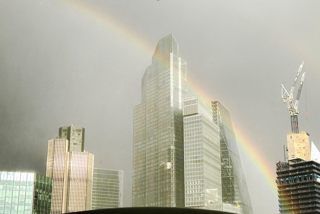 Rainbow arches over modern skyscrapers and crane in a cloudy city skyline, creating a serene urban scene.