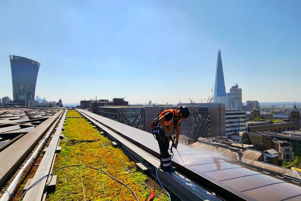 Worker in safety gear cleaning solar panels on rooftop with iconic London skyline backdrop.