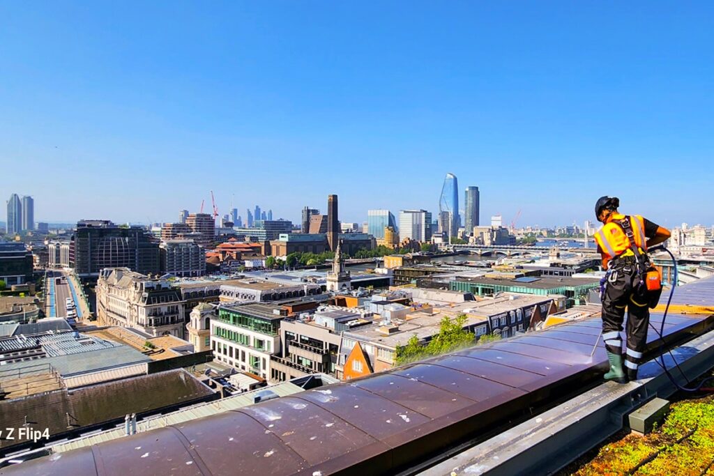 Worker in safety gear cleaning rooftop with hose, overlooking sunny London skyline under clear blue sky.