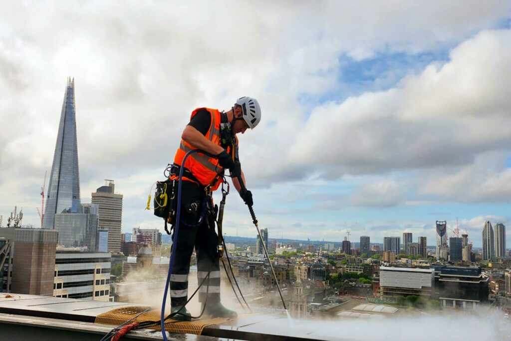 Worker power washing skyscraper rooftop in London, with The Shard in the background under cloudy sky.