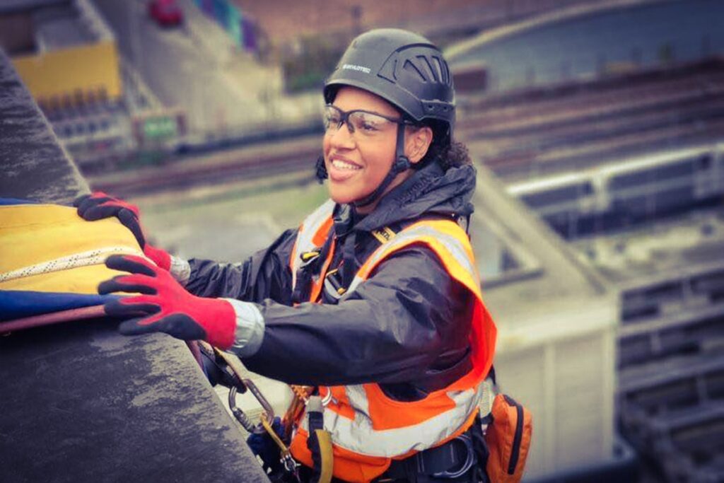 Smiling construction worker in safety gear abseiling on a building, wearing an orange vest and helmet.