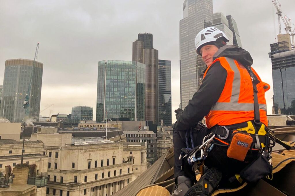 Worker in orange high-vis gear and helmet atop London rooftop, city skyline in cloudy background.