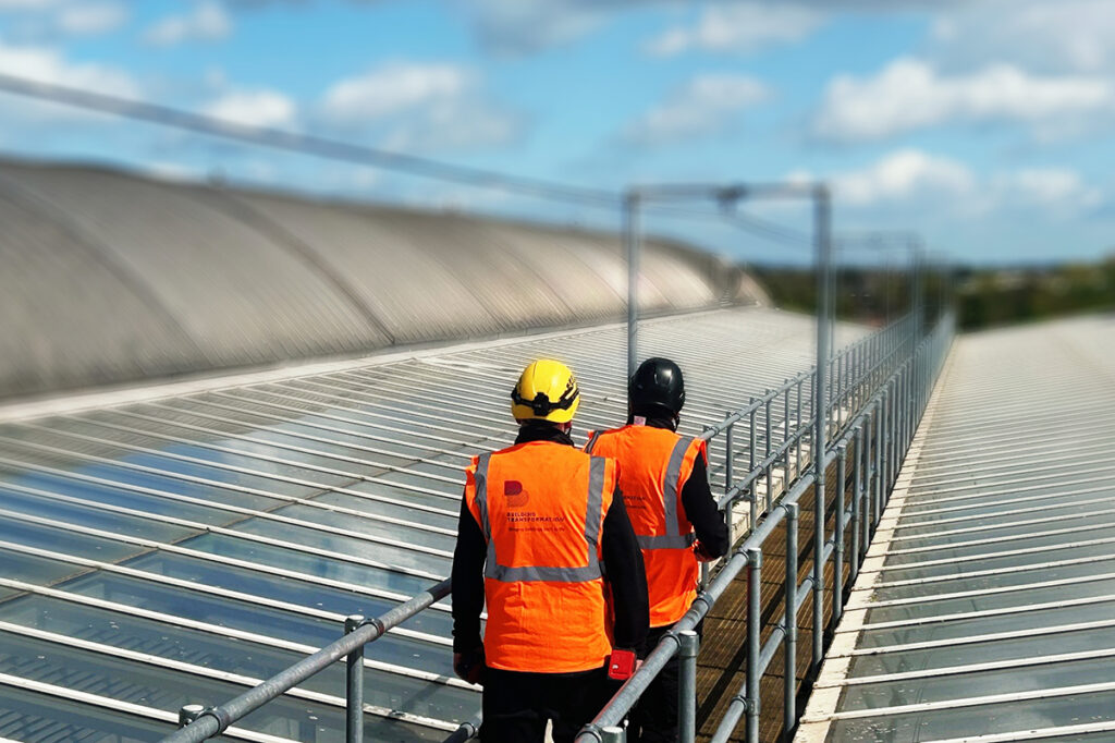 Workers in high-vis vests and helmets on a steel rooftop, under a vivid blue sky with scattered clouds.