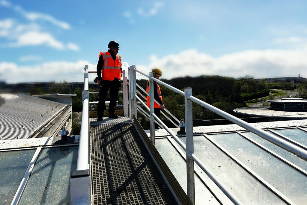 Construction workers in bright orange safety vests inspect a rooftop with metal railings under a clear sky.