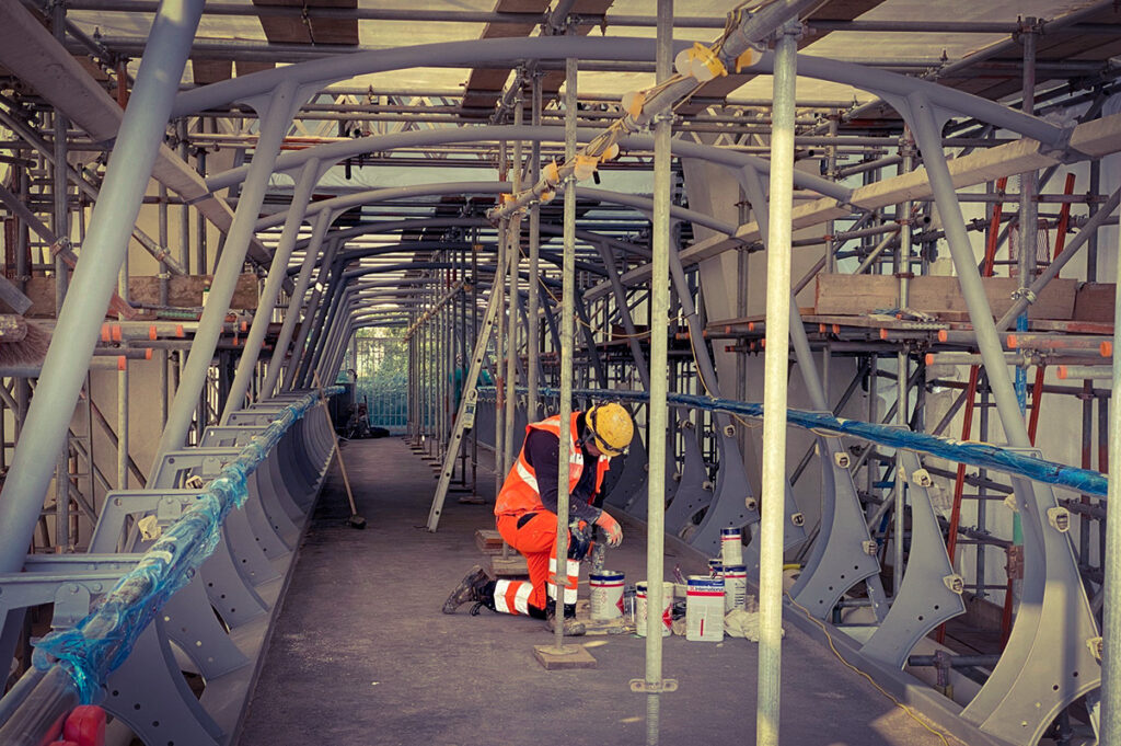 Construction worker in safety gear kneels, painting interior metal structure with scaffolding all around.
