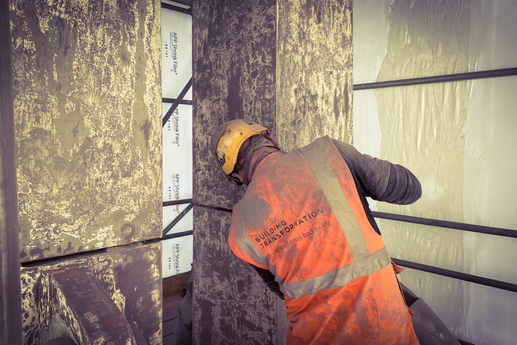 Construction worker in orange vest inspects textured concrete wall. Text reads Building Transformation.