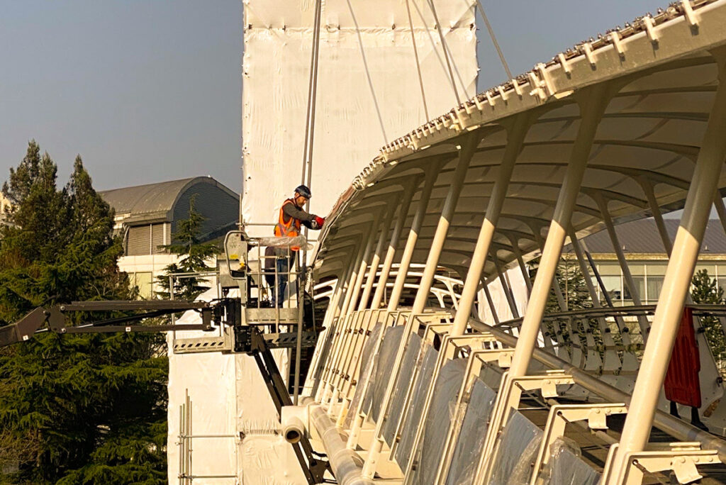 Worker in safety gear operates lift, inspecting modern architecture roof structure under sunny sky.