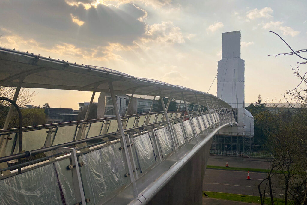 Curved pedestrian bridge during sunset with scaffolding on a tower, under a cloudy sky.