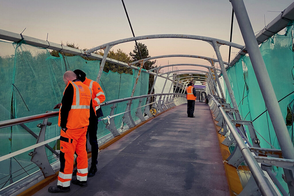 Workers in bright orange vests stand on a modern pedestrian bridge with teal netting against a twilight sky.