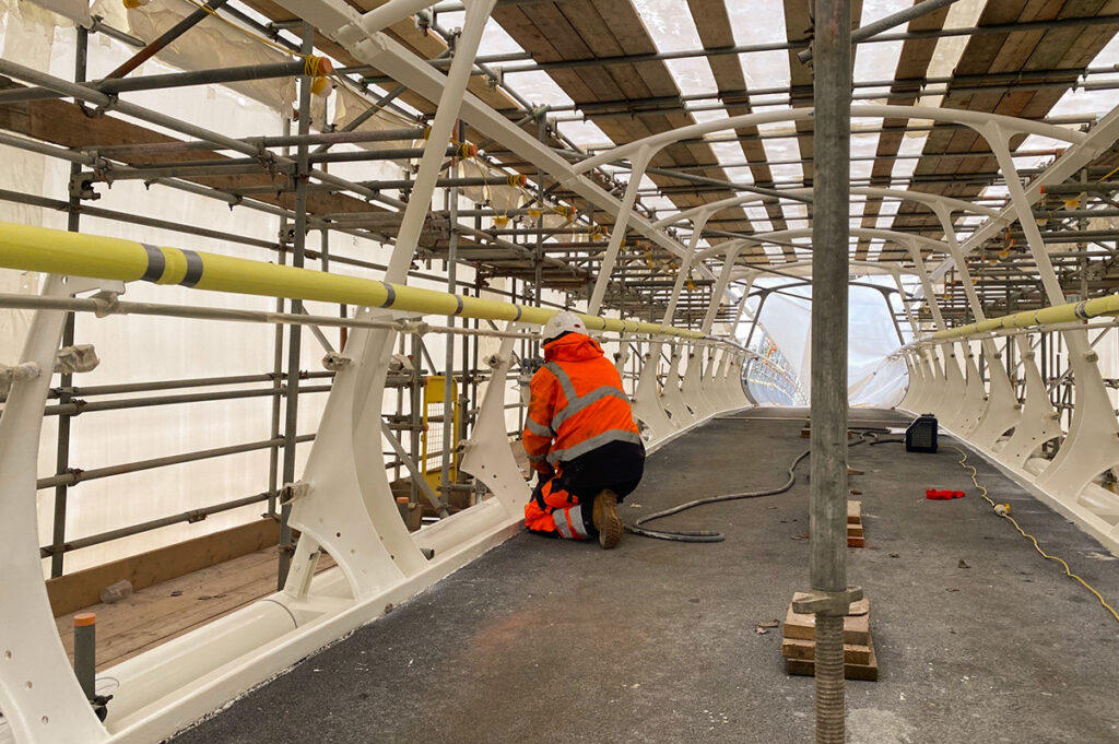 Construction worker in high-vis gear inspects bridge structure within a scaffolded enclosure.