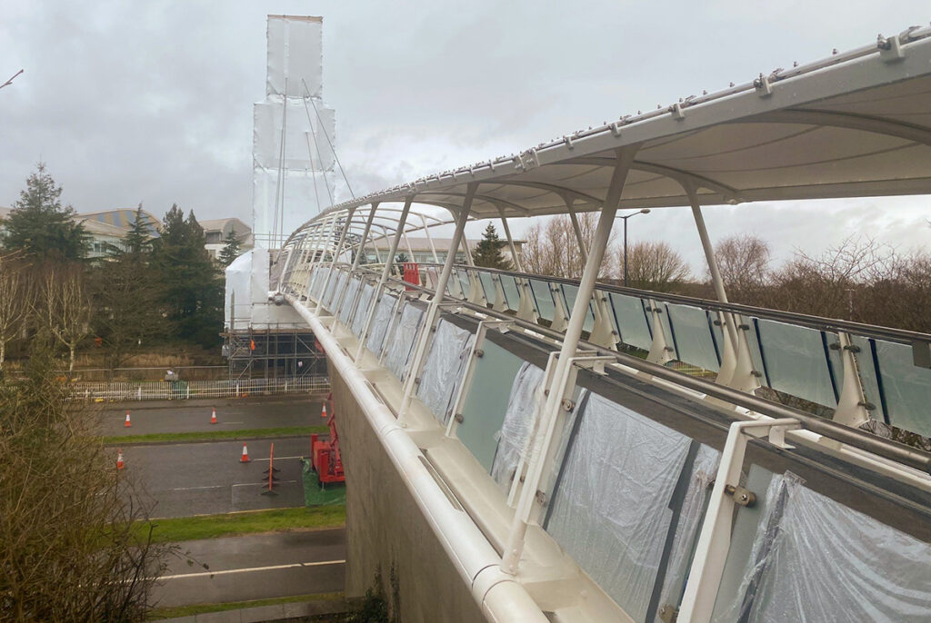Modern footbridge under construction, featuring steel framework and glass panels, set against a cloudy sky.