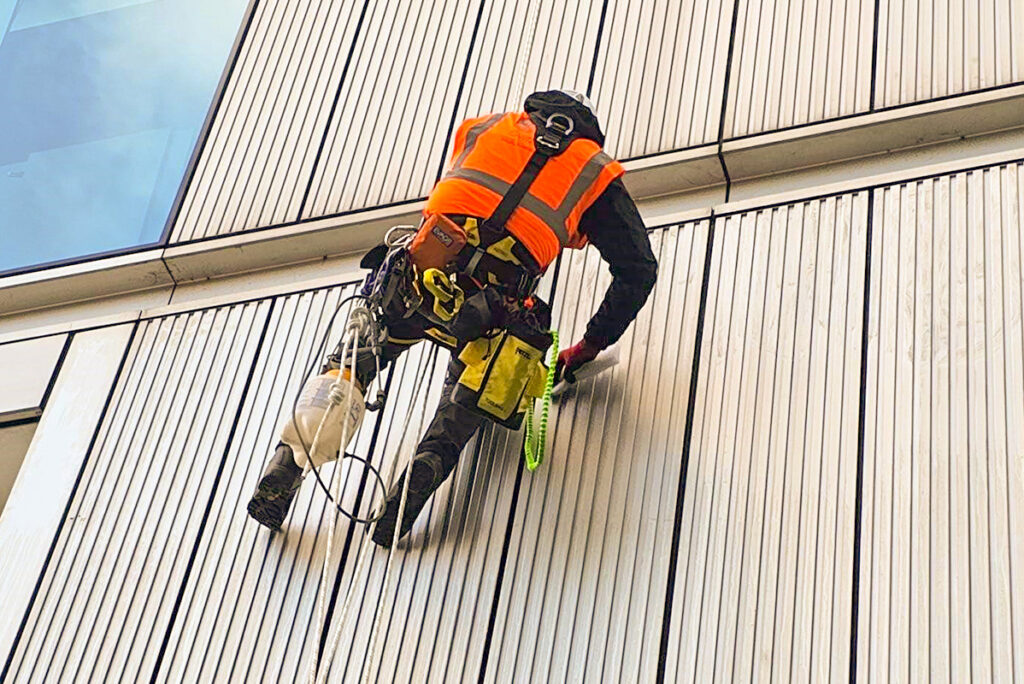 Construction worker in orange vest cleaning high-rise building facade using rope access equipment.