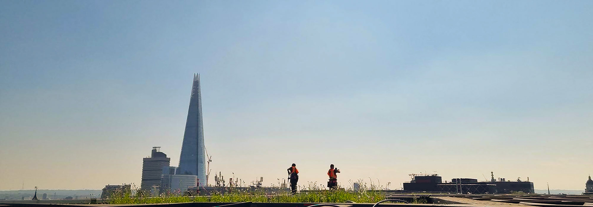 Workers in orange vests on a rooftop with The Shard skyscraper and a clear blue sky in the background.