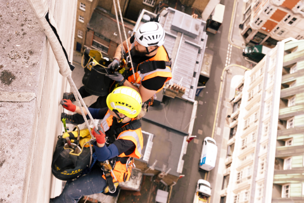 Two workers in helmets and harnesses abseiling a building, wearing orange safety vests, cityscape below.