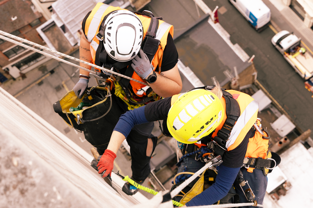 Two construction workers in safety gear abseiling down a building, using ropes and harnesses.