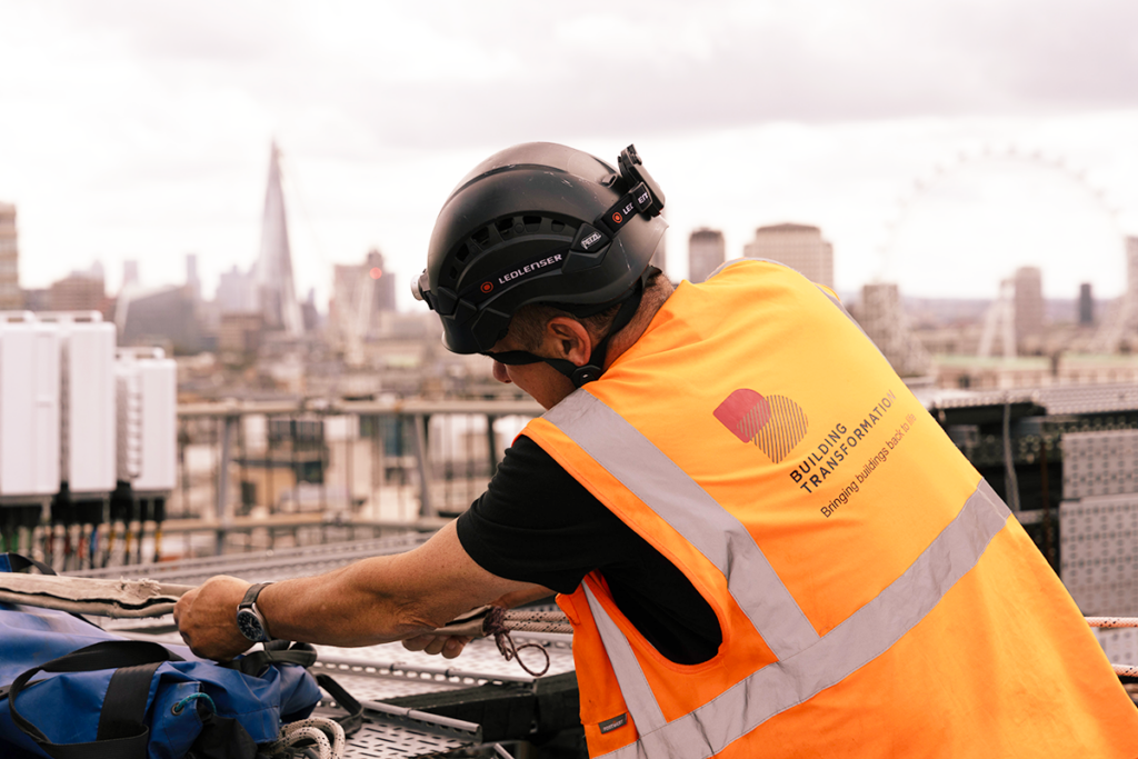 Construction worker in orange vest and helmet from Building Transformation, on rooftop with London skyline.