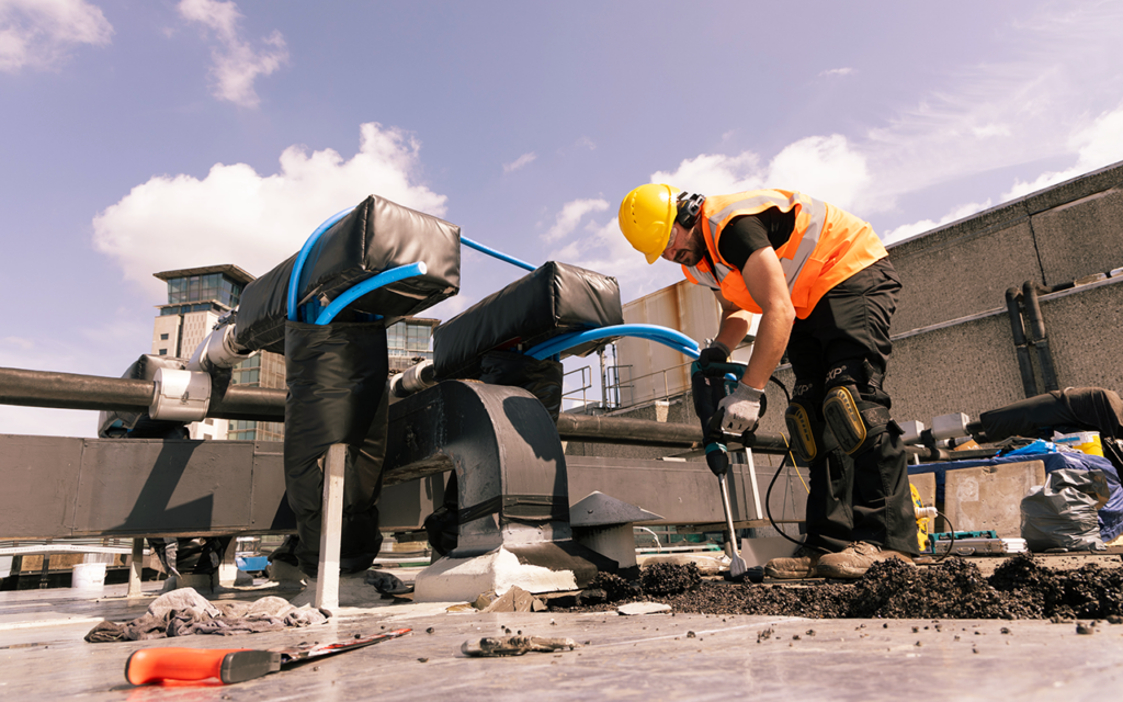 Construction worker drilling on rooftop, wearing high-vis vest and helmet under a bright blue sky.