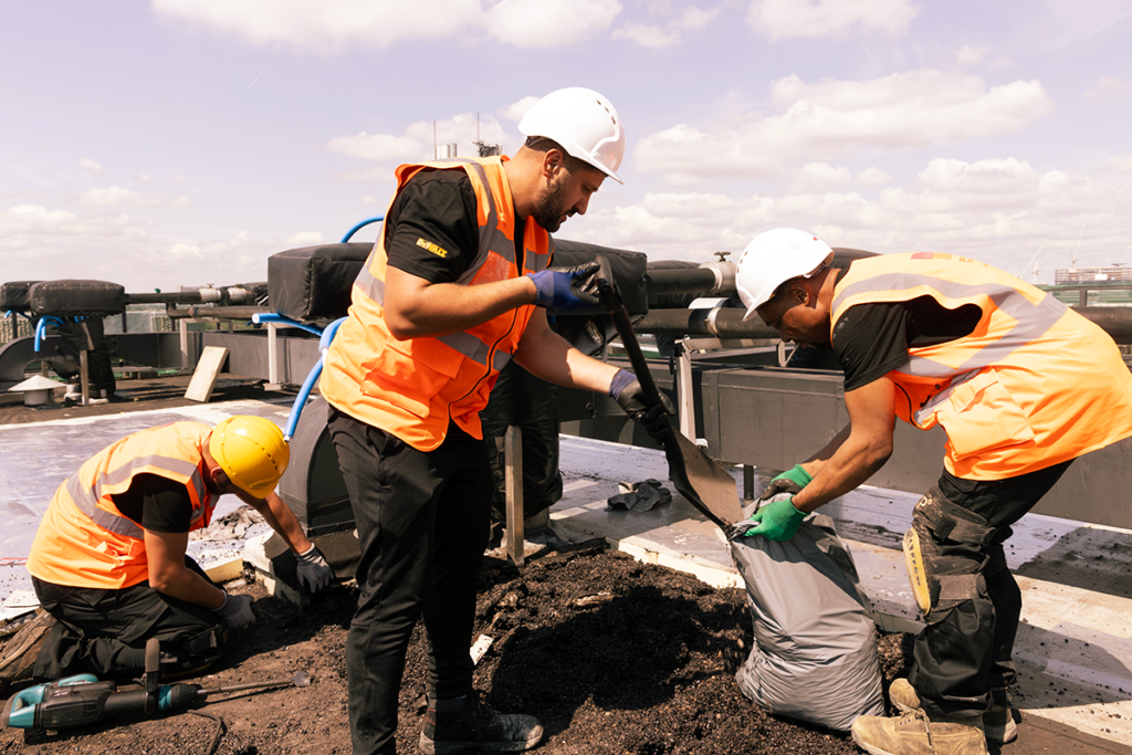 Construction workers in safety gear on a rooftop, shovelling gravel into bags under a sunny sky.