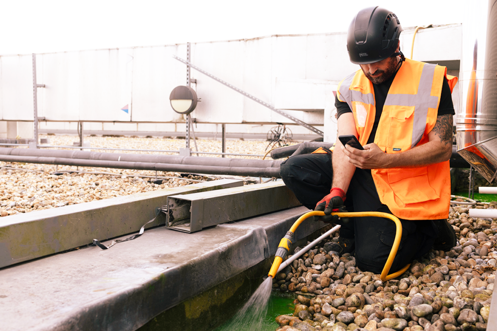 Worker in orange safety vest and helmet using a phone, kneeling with a hose on a pebble-covered rooftop.