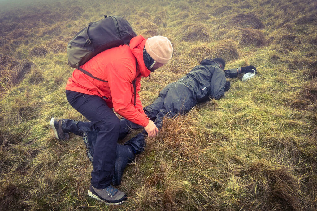 Hiker in red jacket helps fellow hiker on grassy moorland, emphasising outdoor rescue teamwork.