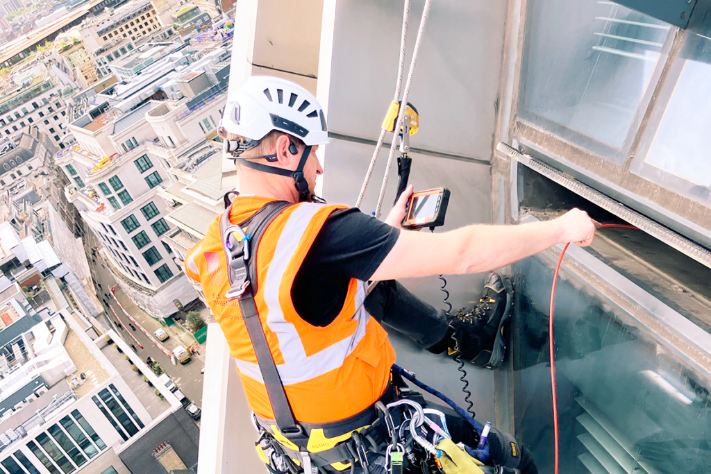 High-rise worker in safety gear and helmet inspecting a building facade with tools overlooking cityscape.