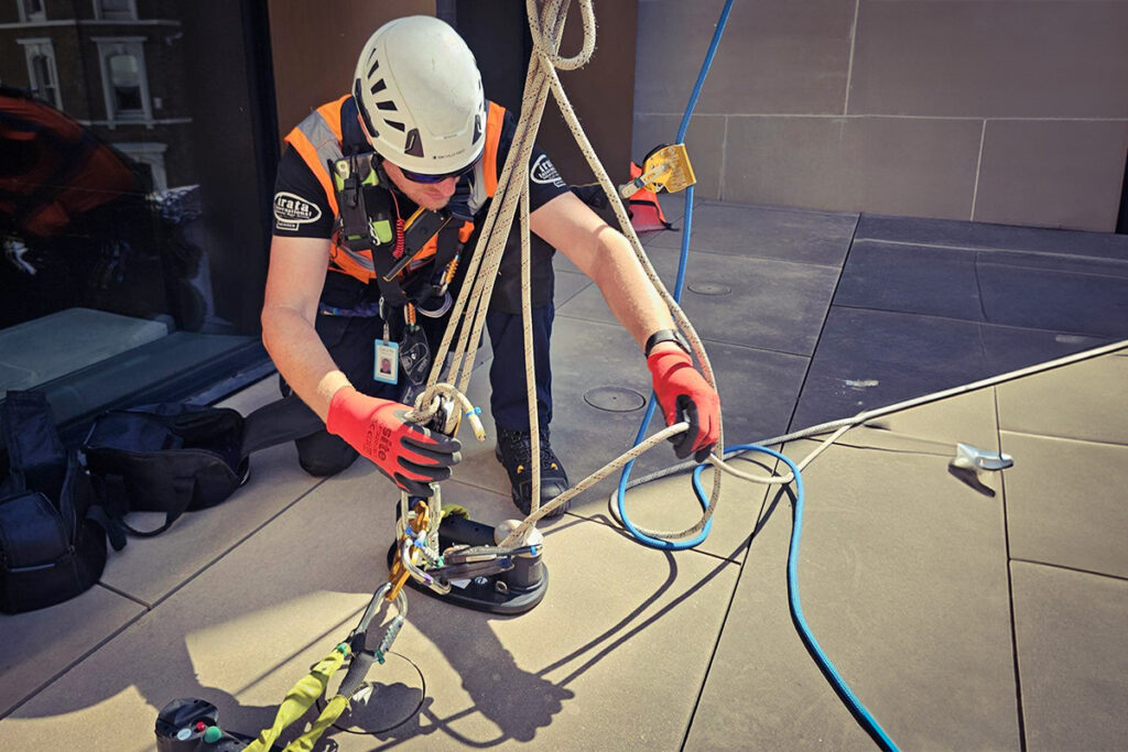 Worker in safety gear adjusts rope system on building rooftop, wearing white helmet and red gloves.