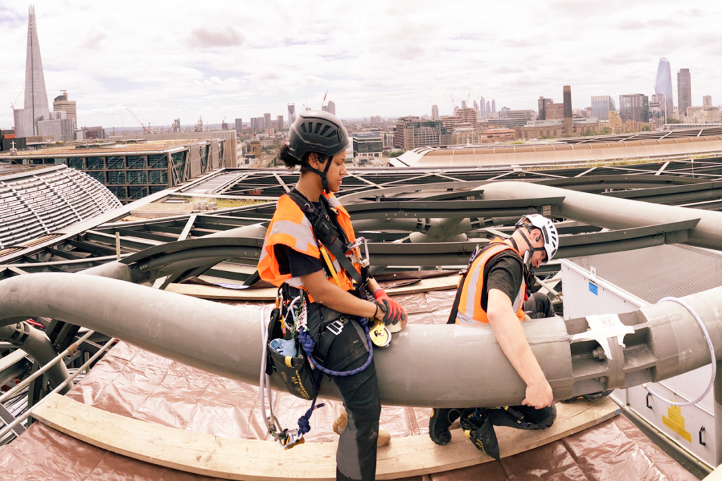 Construction workers in harnesses and helmets, inspecting pipes on a London rooftop with cityscape view.