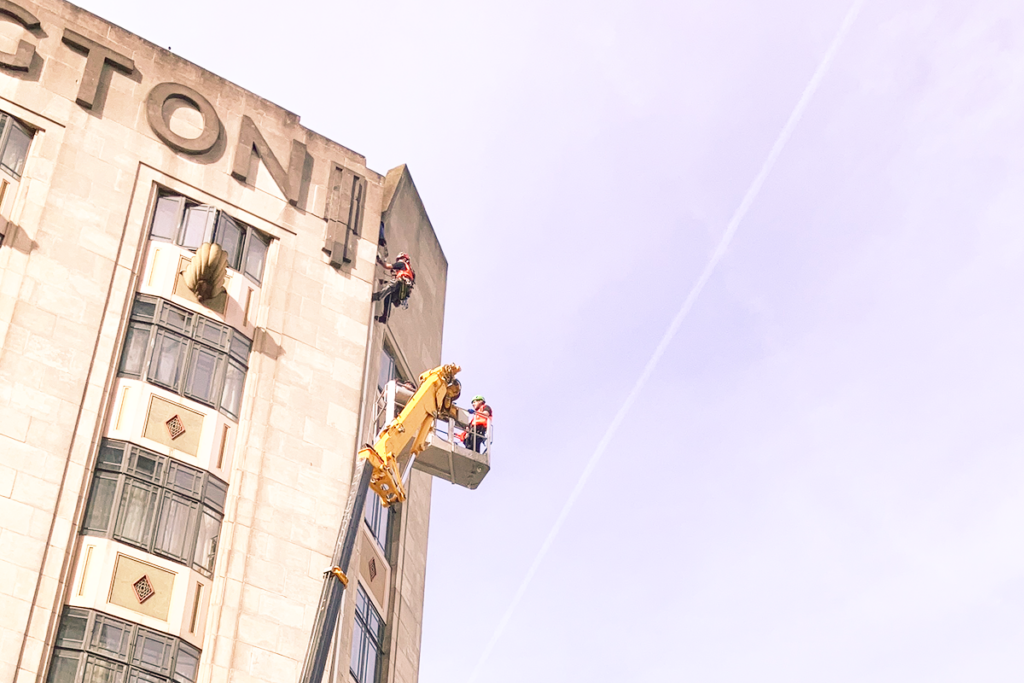 Workers in high-vis gear on crane platform and ropes inspecting art deco building facade under blue sky.