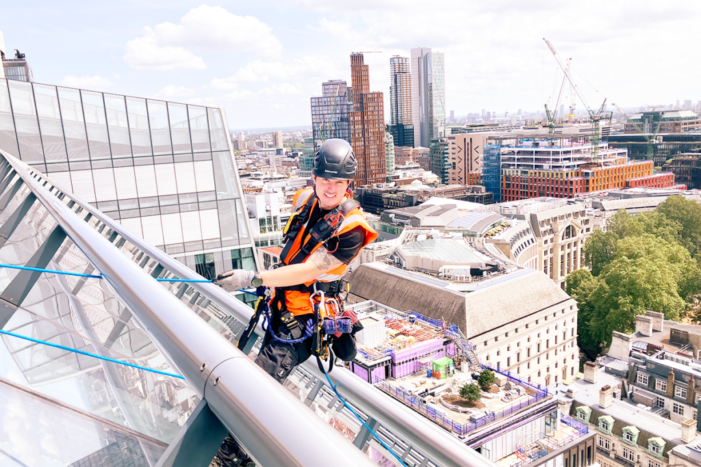 Construction worker in safety gear scaling a skyscraper in a vibrant cityscape with cranes and buildings.