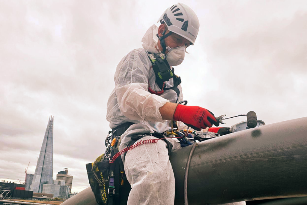 Worker in safety gear painting a pipe on a rooftop with The Shard visible against a cloudy sky.